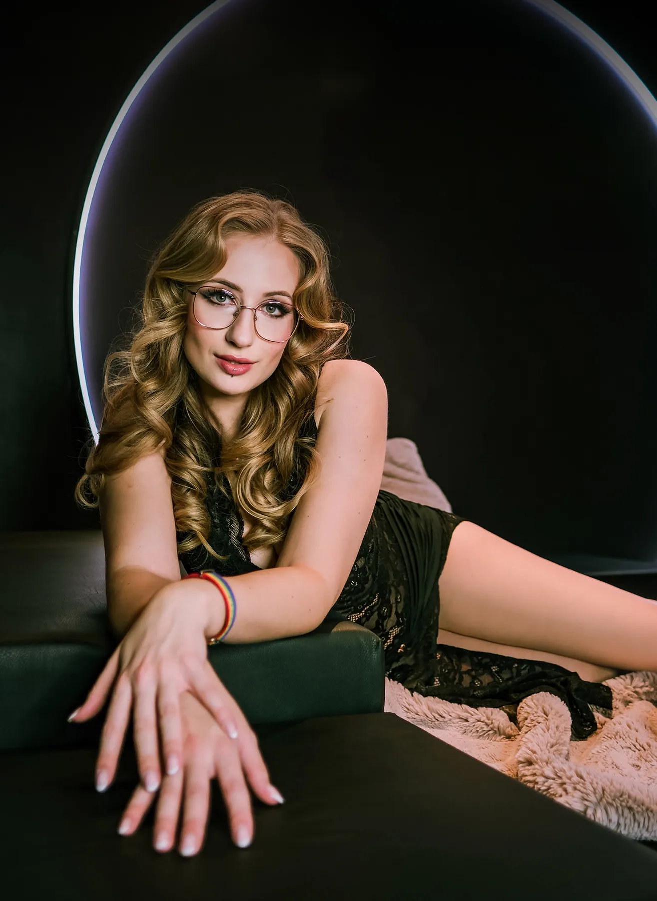Mika Nox mit Brille im schwarzen Spitzen-Body auf einer Ledercouch, Neon-Ring im Hintergrund
